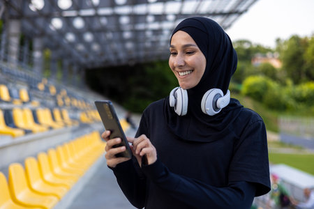 Muslim sportswoman in hijab at stadium with headphones and phone in hands, woman during active exercise and fitness, outdoors listening to online music from audio books and podcasts app.の写真素材