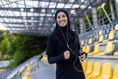Young beautiful Muslim woman running in hijab in stadium, sportswoman smiling in headphones holding phone, listening to audio book.の写真素材