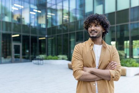 Portrait of an Indian young man, confident businessman, office worker standing outside an office building, arms folded across his chest and looking to the side smiling.の写真素材