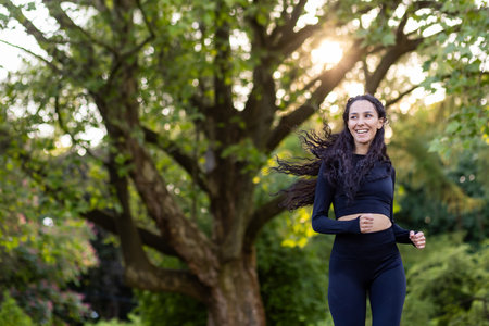 Young motivated and joyful woman jogging in the morning in a public park among trees, Hispanic woman with curly hair, slim in a tracksuit.の写真素材