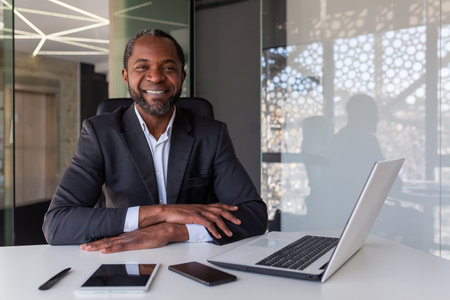 Portrait of successful mature adult businessman, man with crossed arms smiling and looking at camera, african american boss financier sitting at table with laptop.の写真素材