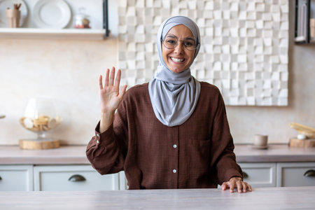 Muslim young woman in hijab sitting in the kitchen at home and smiling talking and greeting to the camera.の写真素材