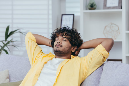Young man relaxing alone at home on sofa, close-up Hispanic man with hands behind head dozing and dreaming with closed eyes in living room of house.の写真素材