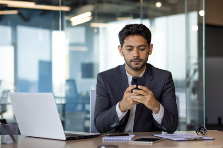 Serious thinking businessman using phone at workplace, arab boss in business suit thinking inside office reading online news using app on smartphone.の写真素材