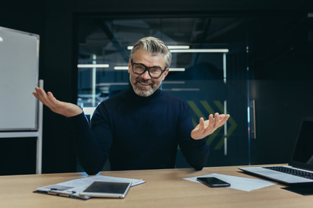 Portrait of an older male businessman sitting in the office smiling and talking on a video call, conducting a webinar, an online business meeting.の写真素材