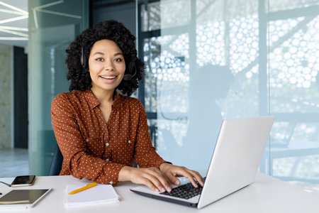 Portrait of female office worker with video call headset and laptop inside office at workplace,...