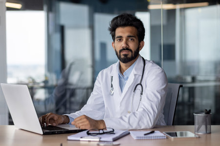 Portrait of a young doctor student studying online at a laptop, sitting in the office in a white coat. He looks seriously into the camera.の写真素材