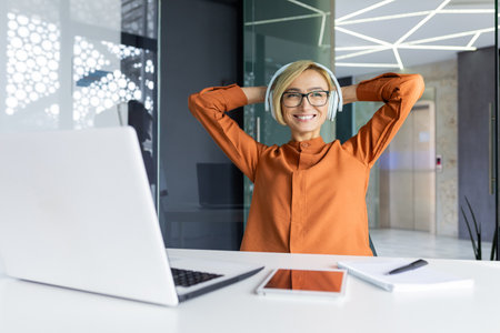 Young successful and satisfied business woman at the workplace completed a work project, female worker with a laptop and headphones has her hands behind her head, is smiling, listening to music.の写真素材