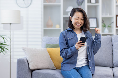 Woman celebrating victory at home looking at phone screen, satisfied asian woman happy received online notification of good achievement results sitting on sofa in living room.の写真素材