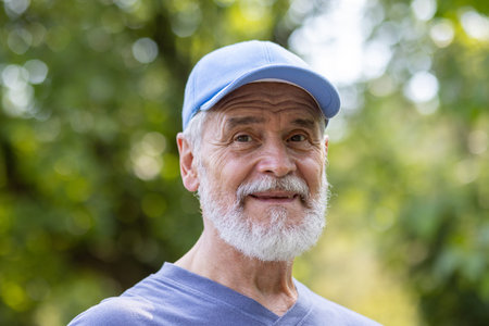 Portrait of senior mature gray-haired man in park, pensioner in sports cap smiling and looking at...
