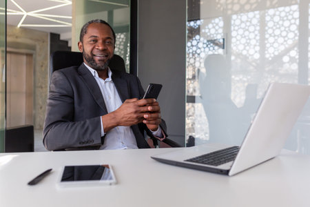 Portrait of senior gray haired african american boss, man smiling and looking at camera at workplace inside office, businessman holding phone, using online app on smartphone.の写真素材