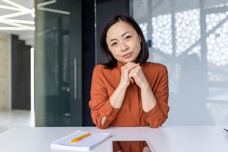 Portrait of serious confident business woman, Asian looking thoughtfully at camera, listening to conversation and report, video ringing online meeting with colleagues remote inside office.の写真素材