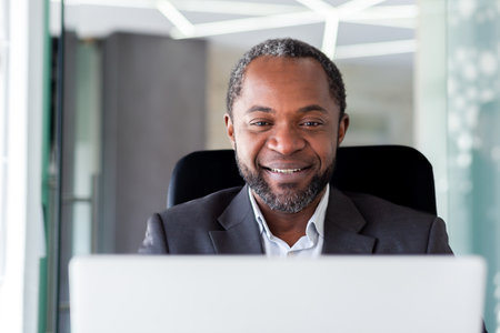 Mature adult african american businessman in middle of office working with laptop, close up man smiling looking at computer screen, satisfied with achievements boss at workplace close up.の写真素材