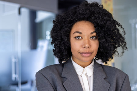 Close up portrait of serious confident business woman, african american woman with curly hair and in business suit looking at camera, female worker inside office at workplace.の写真素材