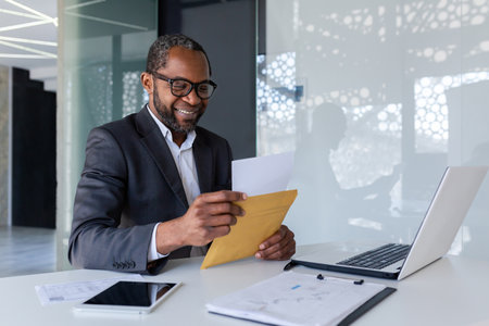 Senior satisfied financier received a letter mail message in an envelope, businessman reads and smiles, boss at desk inside office uses laptop.の写真素材