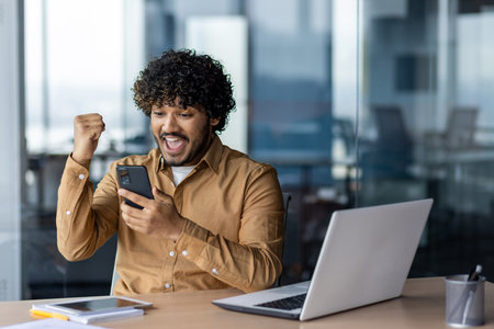 Portrait of hispanic winner at workplace, businessman received online win notification on phone app, man celebrating victory joyfully holding hand up, inside office with working with laptop.の写真素材