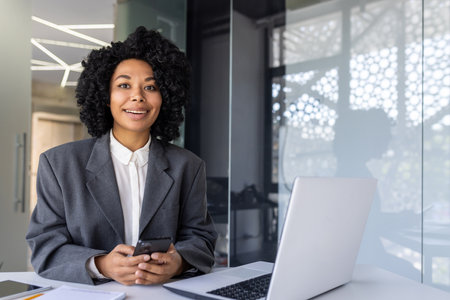 Portrait of a young successful African-American businesswoman sitting at a desk in an office at a desk, confidently looking at the camera, holding a phone in her hands.の写真素材