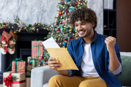 A happy young Indian man is happy to receive news in the mail on Christmas holidays. Sitting at home on the sofa and reading a letter in an envelope, showing a victory gesture with his hand yes.の写真素材