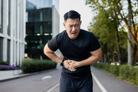 Asian young male athlete bent over from pain in his side, holding his stomach with his hands after...