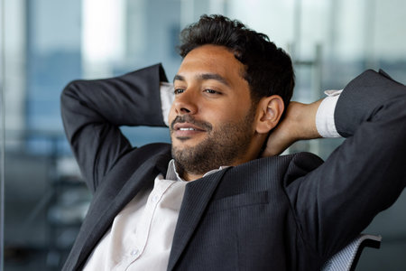 Dreamy thinking man inside office close up smiling and looking out the window, businessman in business suit with hands behind head resting and visualizing future results of achievement and victory.の写真素材