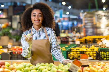 Portrait of a young saleswoman in a supermarket, grocery store, a smiling saleswoman looking at the camera with satisfaction, looking at apples, a Hispanic woman with curly hair in an apron.の写真素材