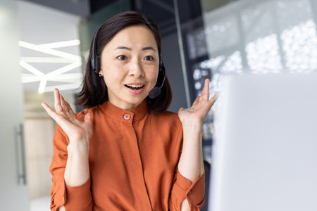 Surprised joyful and smiling woman looking at laptop screen, businesswoman using headset phone, at workplace for video call.の写真素材