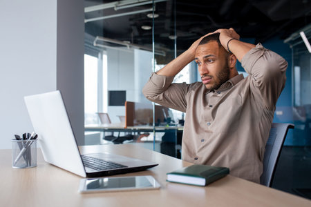 Frustrated and sad man at workplace reading online message from laptop, businessman sitting inside office at workplace, desperate unhappy, got bad news, bankruptcy, layoff.の写真素材