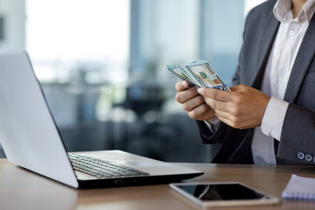Close-up portrait of male hands counting dollars, man sitting at desk in office using laptop in suit and counting money earned.の写真素材
