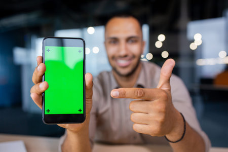 Smiling man holding a phone with a green screen in his hands, showing it to the camera and pointing...