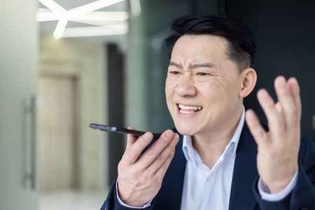 Close-up photo of an angry and serious Asian male businessman talking emotionally on the phone through a loudspeaker in the office.の写真素材