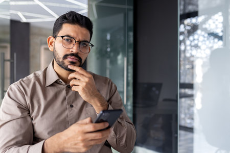 Portrait of successful Indian businessman, serious thinking man looking at camera at workplace inside office, boss holding phone, using laptop.の写真素材