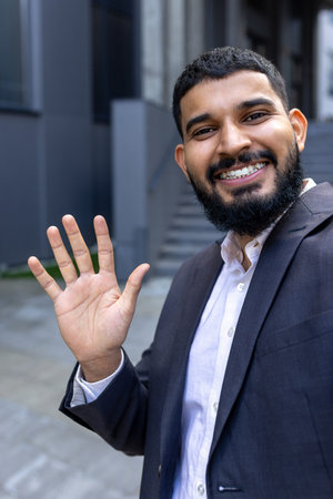 Vertical close-up photo of a young smiling Muslim businessman man talking on the phone and waving at...