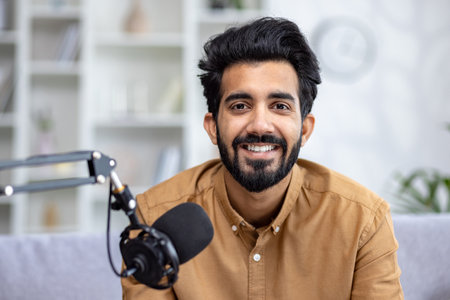 Close-up portrait of a young Indian man sitting at home on the couch in front of a microphone stand and smiling at the camera.の写真素材