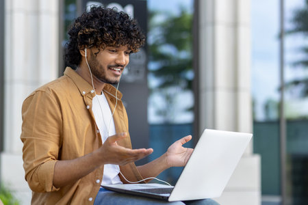 Indian young man sitting outside on a bench with a laptop and headphones and talking online on a video call.の写真素材