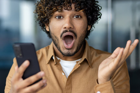 Close-up portrait of a young Indian man looking shocked and surprised at the camera while holding a mobile phone and spreading his hands.の写真素材