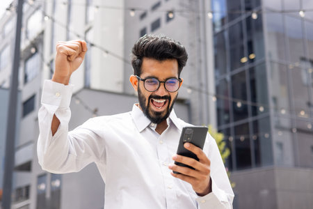 Young successful hispanic man in casual clothes outside office building received online win notification, man celebrating victory triumph, using app on phone, good news in businessmanの写真素材