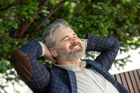 Mature businessman in suit enjoys a break outdoors, smiling with hands behind head, trees in the background.の写真素材