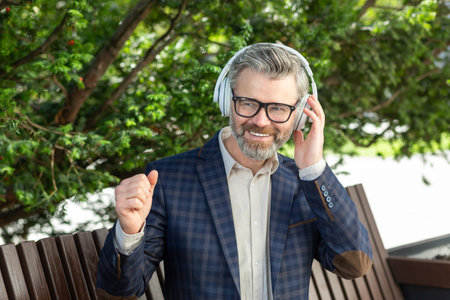 A smiling senior businessman in a stylish suit relaxes with music on headphones while seated outdoors, exuding confidence.の写真素材