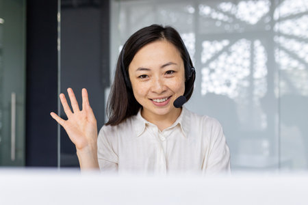 Professional Asian businesswoman in office with headset smiling and waving hand, symbolizing approachability and customer service.の写真素材