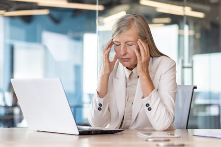 Tired senior gray-haired businesswoman sitting at the desk in the office and holding her head with her hands, feeling pain and tension.の写真素材