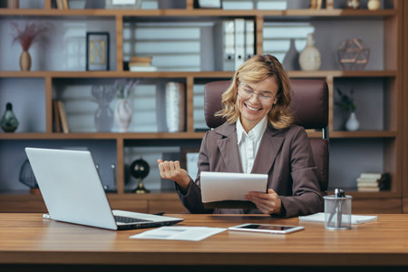 Smiling senior businesswoman in a well-appointed home office, confidently managing remote work with digital tablet and laptop.の写真素材