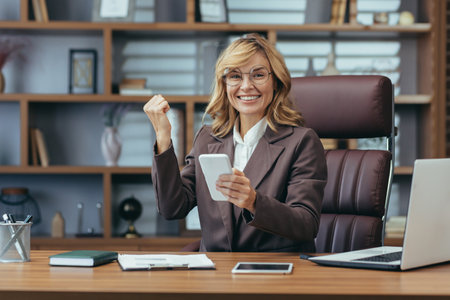 Elegant mature professional blonde woman expressing joy while working remotely in a stylish home office ambiance.の写真素材