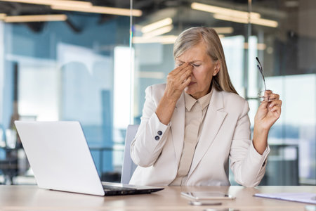 Tired senior woman dressed in office suit sitting at desk with laptop and touching forehead while having headache. Female director taking off glasses to let eyes rest during working at computer.の写真素材
