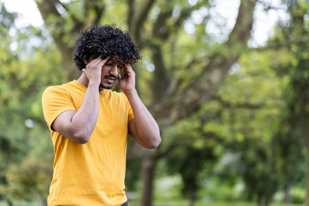 Close-up photo of a young Indian sportsman standing in the park and holding his head with his hands,...