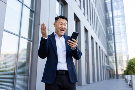Happy young Asian man businessman in suit standing near office building with phone and shocked happy success, spreading arms.の写真素材