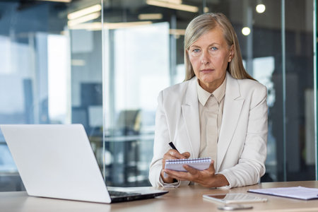 Concentrated senior woman looking at camera with focused sigh while holding notebook and pen by...