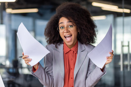 Cheerful young businesswoman with curly hair feeling excited while holding papers in a contemporary...