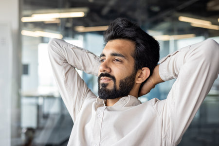 A man with facial hair - including a beard - sits in an office, relaxing with his hands behind his head as a gesture of relaxation.の写真素材