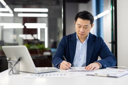Serious thinking man behind paper work inside office, mature asian man in business suit filling data in table, writing with pen, businessman boss at workplace with laptop.の写真素材