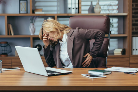 Senior business woman in a suit sitting at a desk in the office holding her hand on her back and head and suffering from severe pain. Needs rest and medical assistance.の写真素材